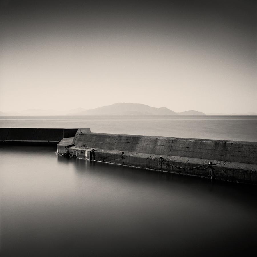 A concrete pier extends into calm water with mountains in the distance under a bright sky.
