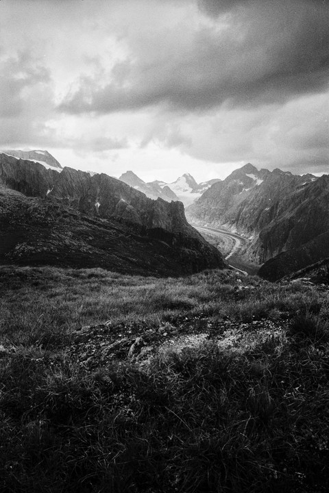 A dramatic black and white landscape shows a valley with a winding glacier surrounded by towering, rocky mountains under a cloudy sky.