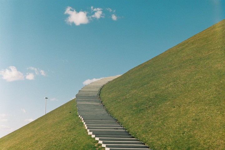 A long concrete stairway curves up a grassy hill towards a clear blue sky with a few white clouds.