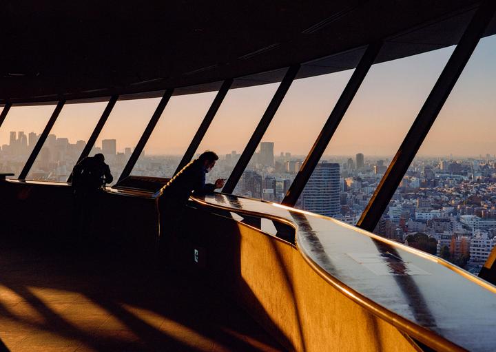 Two men are silhouetted against a cityscape at sunset, viewed from a high vantage point through large windows.