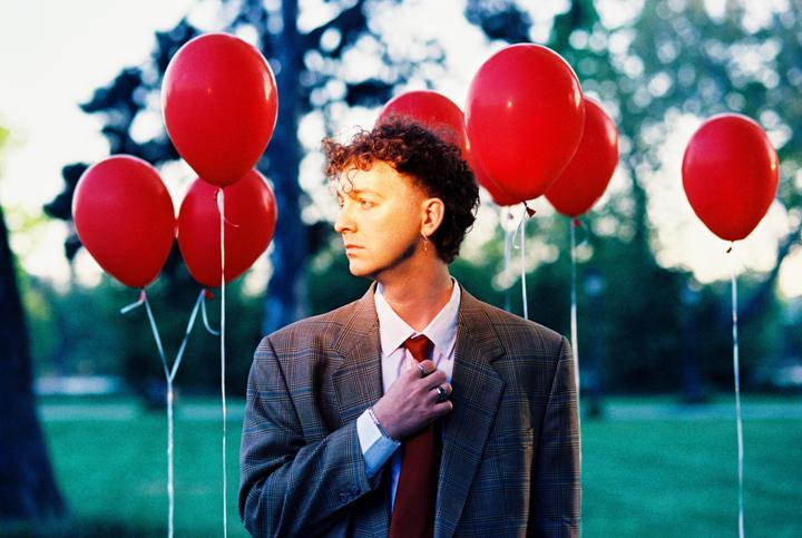 A man in a suit and tie stands among red balloons in a park.
