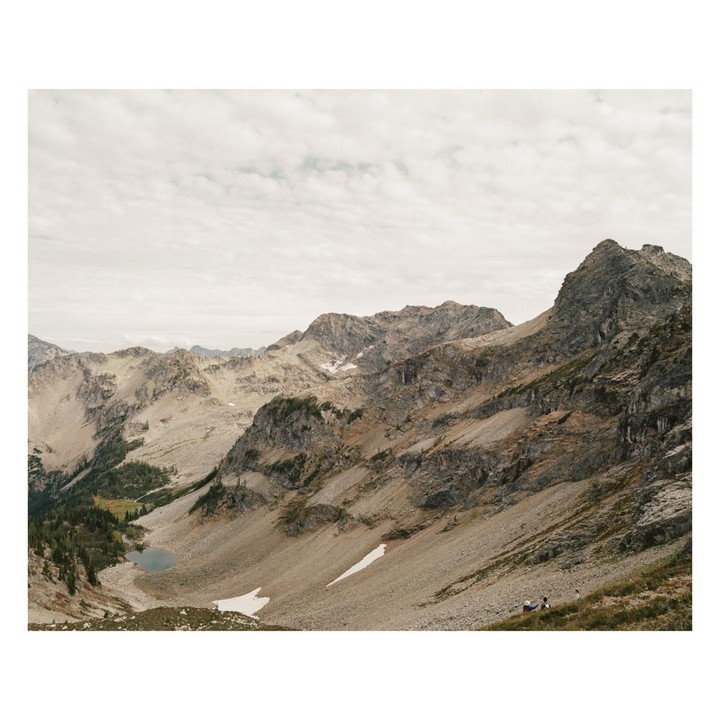 A vast mountain landscape shows rocky slopes, patches of snow, a small lake, and a cloudy sky.