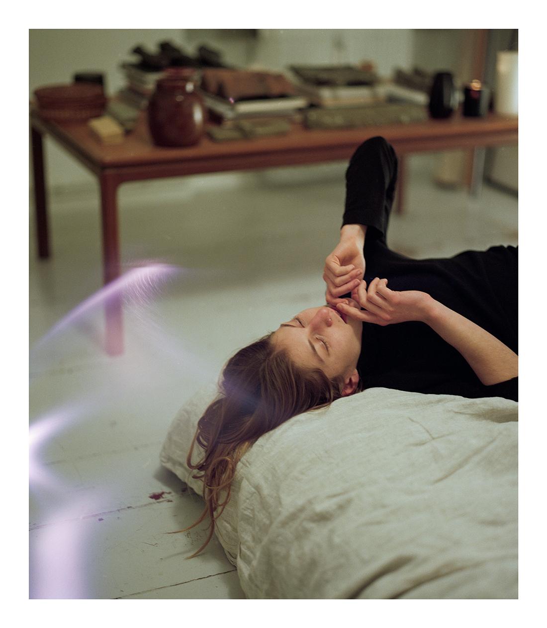A woman lies on a pillow on the floor with her eyes closed and her hands near her mouth, with a table and objects in the background.