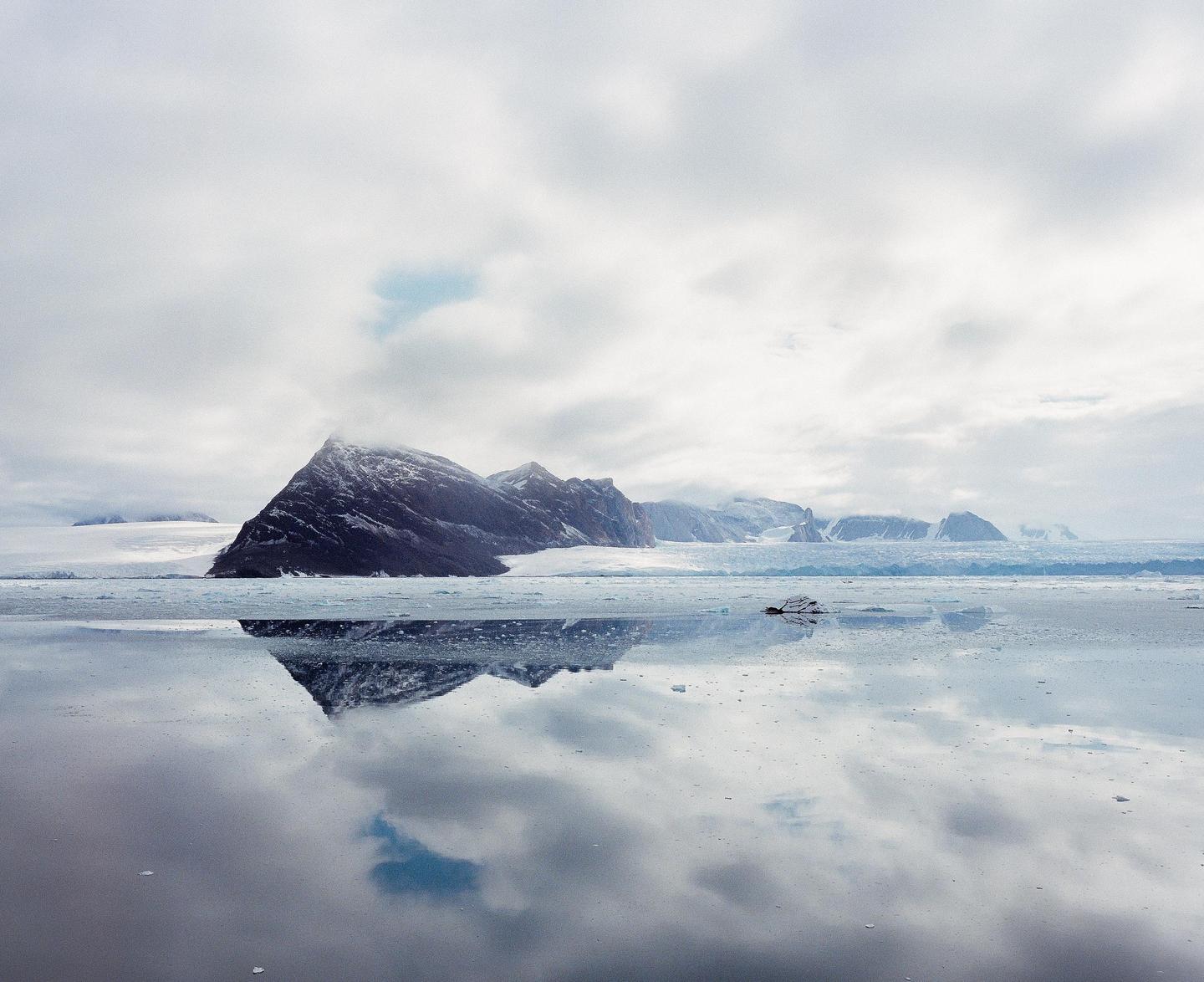A snow-capped mountain range is reflected in a body of water under a cloudy sky.