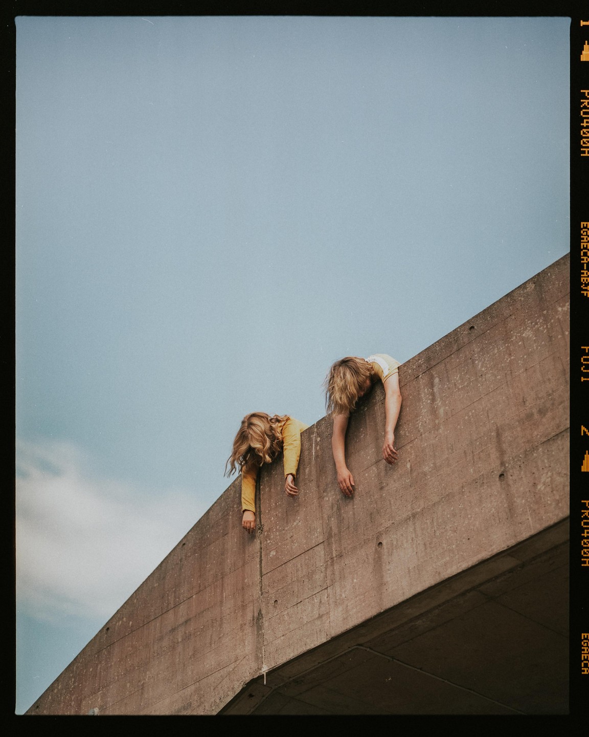 Two children with long hair hang over the edge of a concrete wall against a blue sky.
