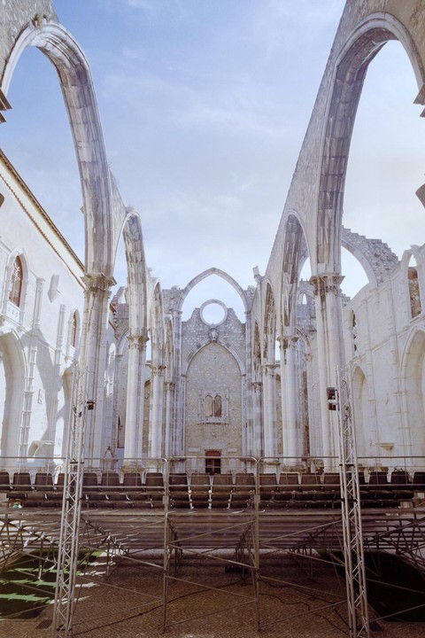 A large, ruined stone building with arched openings and a temporary seating structure in the foreground is shown under a bright sky.