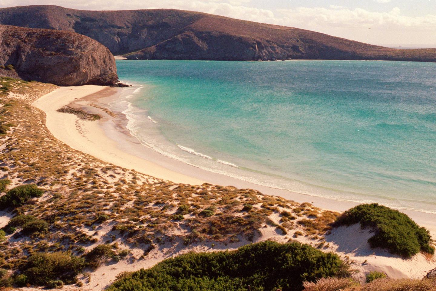 A sandy beach with turquoise water is nestled between a large rock formation and a hill covered in dry grass.