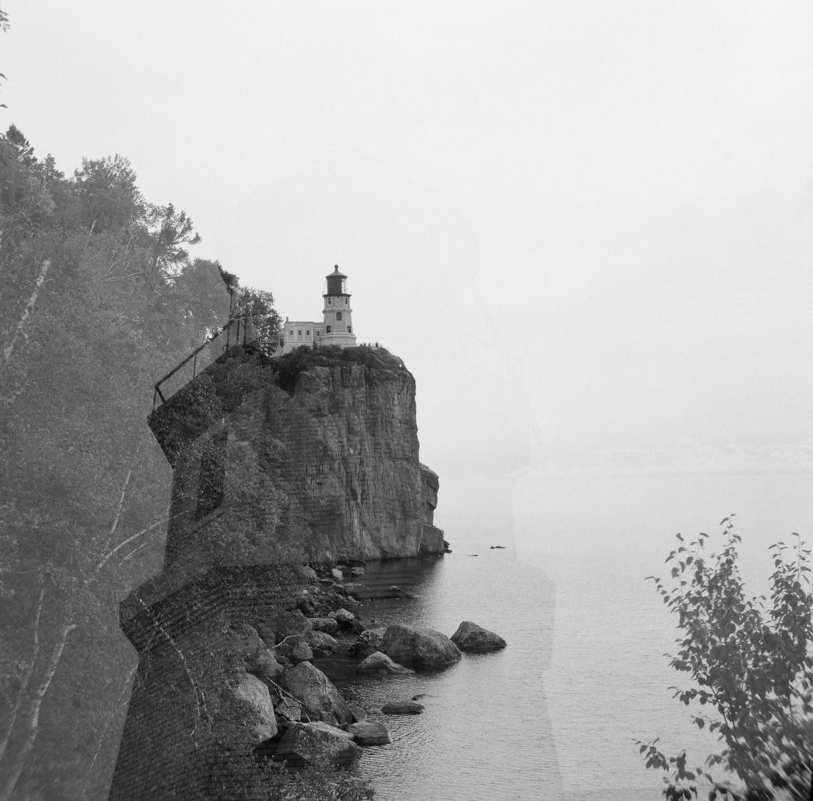 A lighthouse stands atop a cliff overlooking a body of water with trees on the left.