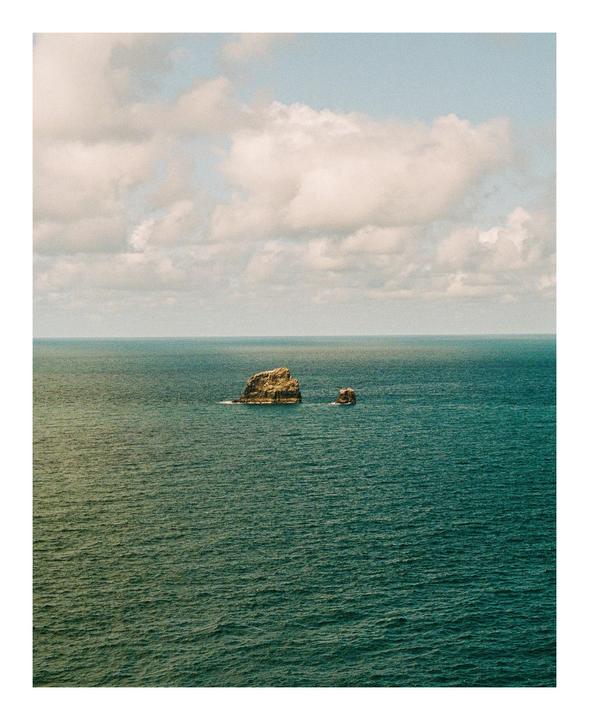 Two small rock formations emerge from the vast expanse of the ocean under a cloudy sky.