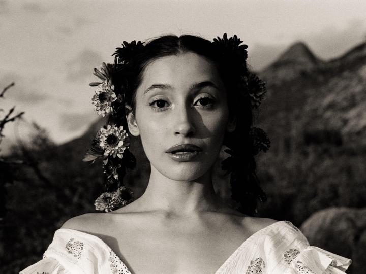 A woman with flowers in her hair and an embroidered top looks directly at the viewer with mountains in the background.