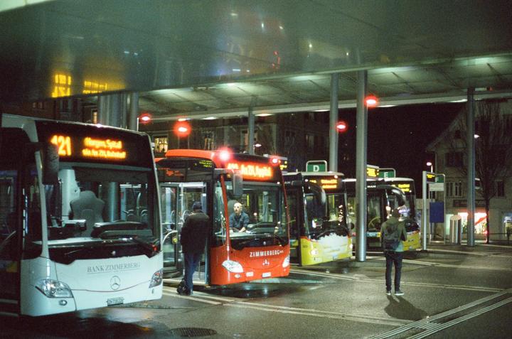 Several buses are parked at a bus stop at night, with people standing nearby.