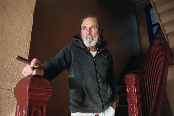 A man with a long beard and hair stands in a stairwell, holding a cigar.