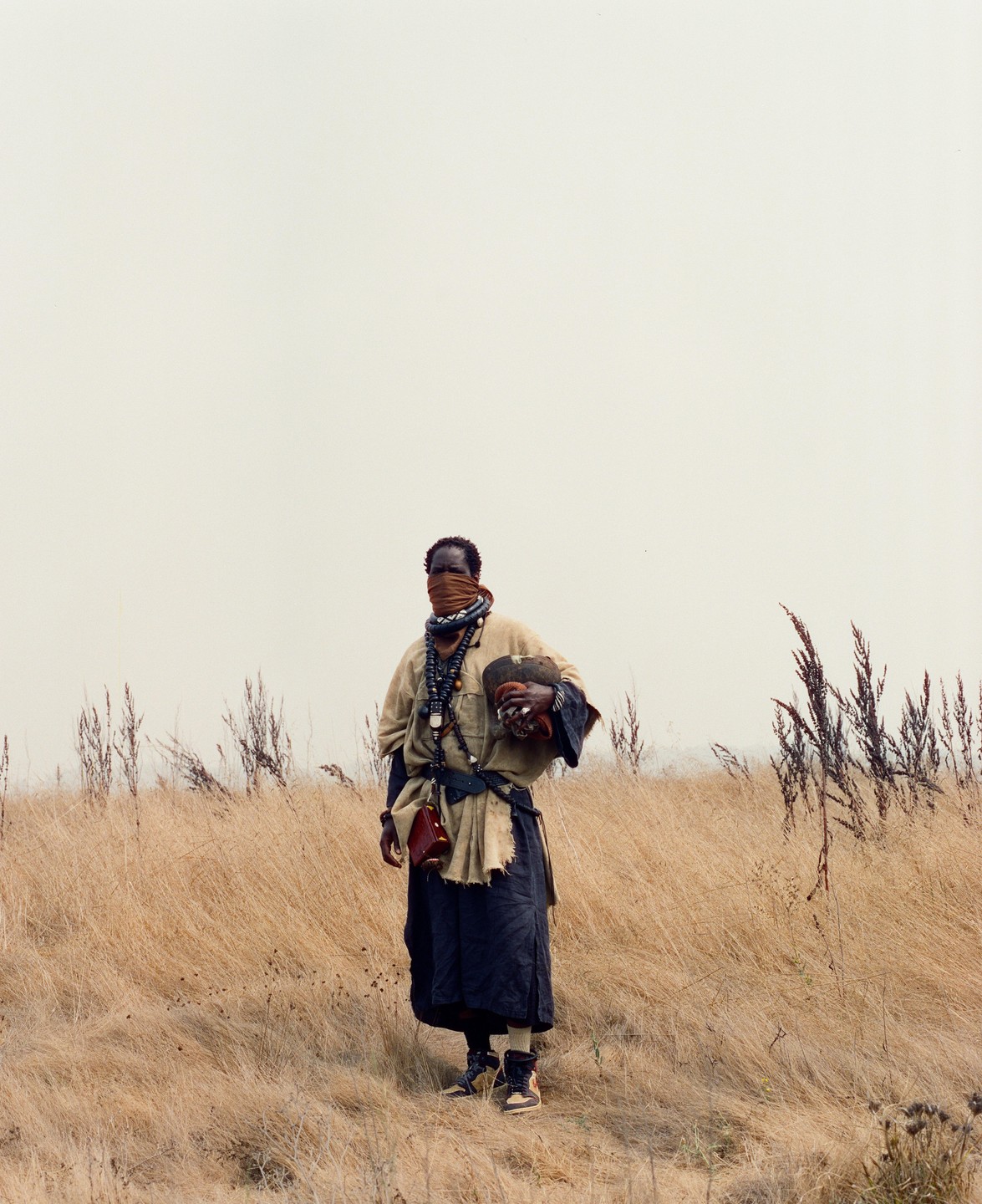A man with a covered face stands in a field of dry grass under a bright sky.