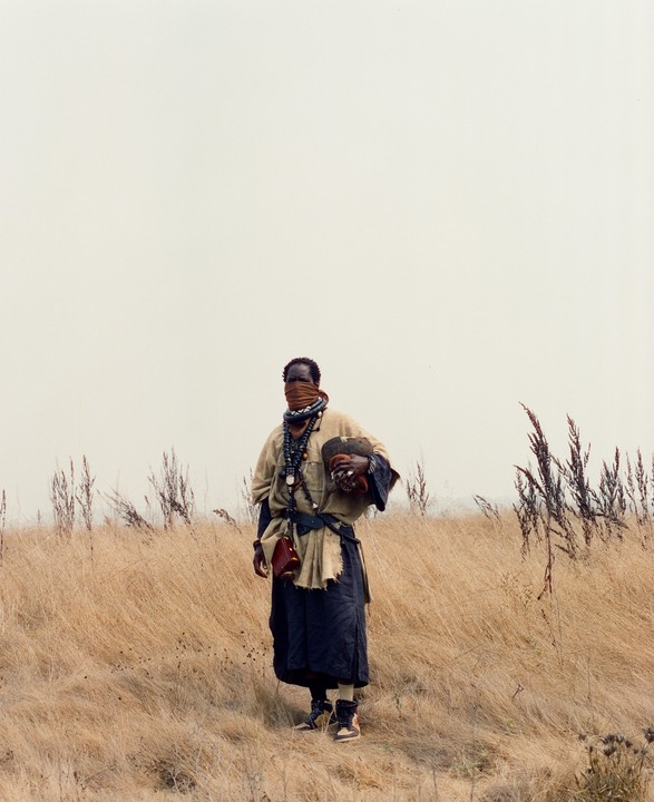 A man with a covered face stands in a field of dry grass under a bright sky.