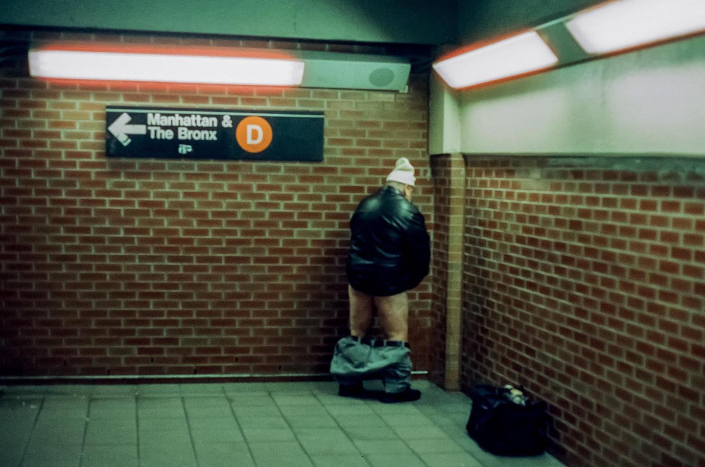 A man with his pants around his ankles stands facing a brick wall in a subway station next to a sign for Manhattan and The Bronx.