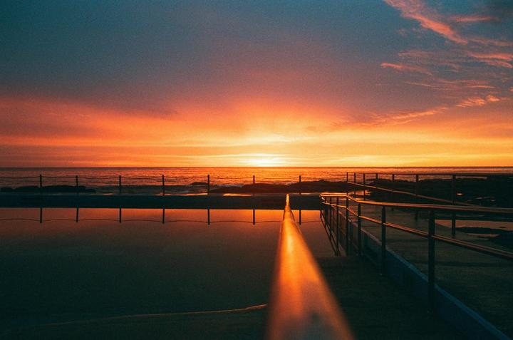 A vibrant sunset illuminates the sky and reflects on the water of a pool next to the ocean.