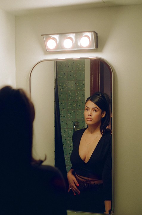 A woman wearing a black top and a cross necklace looks at her reflection in a mirror.