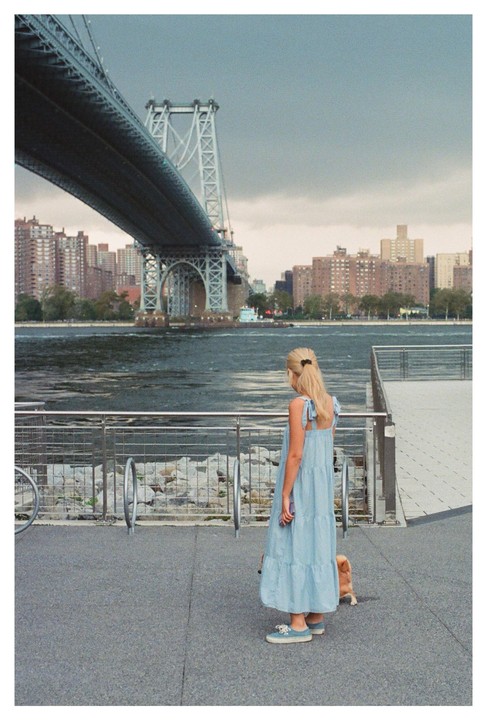 A woman in a blue dress stands on a waterfront promenade with a small dog, looking out at a river and a large bridge.