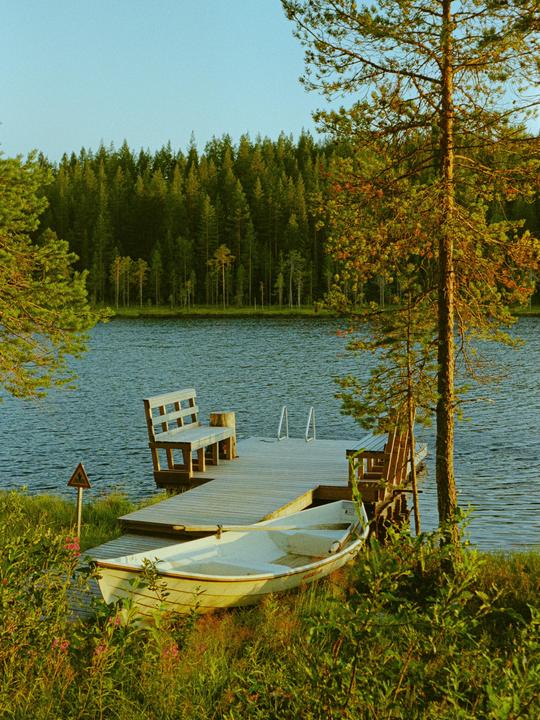 A small boat rests on the grassy shore next to a wooden dock with benches, facing a lake surrounded by a dense forest.