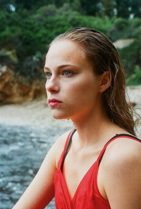 A young woman with wet hair and water droplets on her skin looks to the side, with a body of water and green foliage in the background.