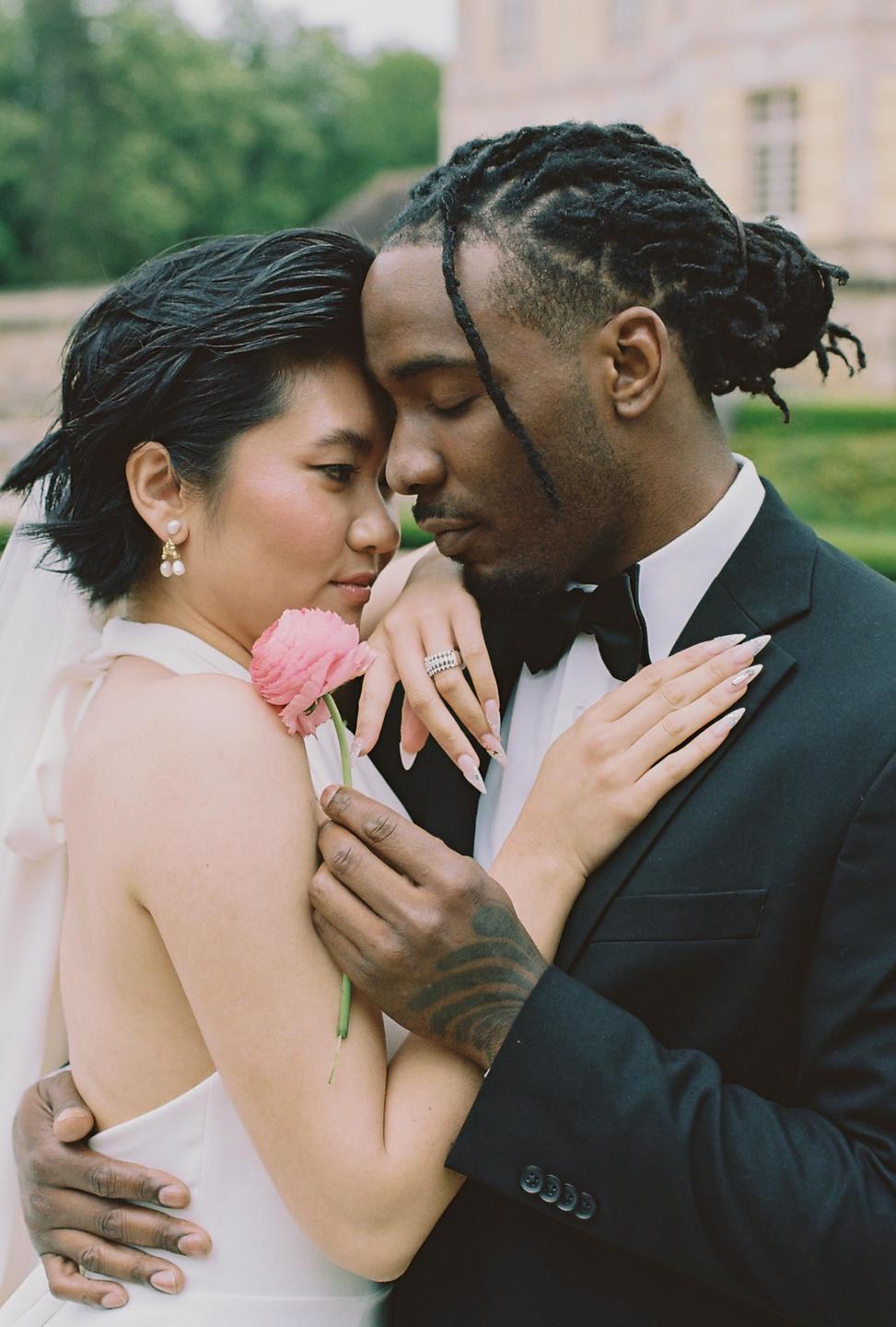 A couple embraces, with the woman holding a pink flower and resting her head on the man's shoulder.