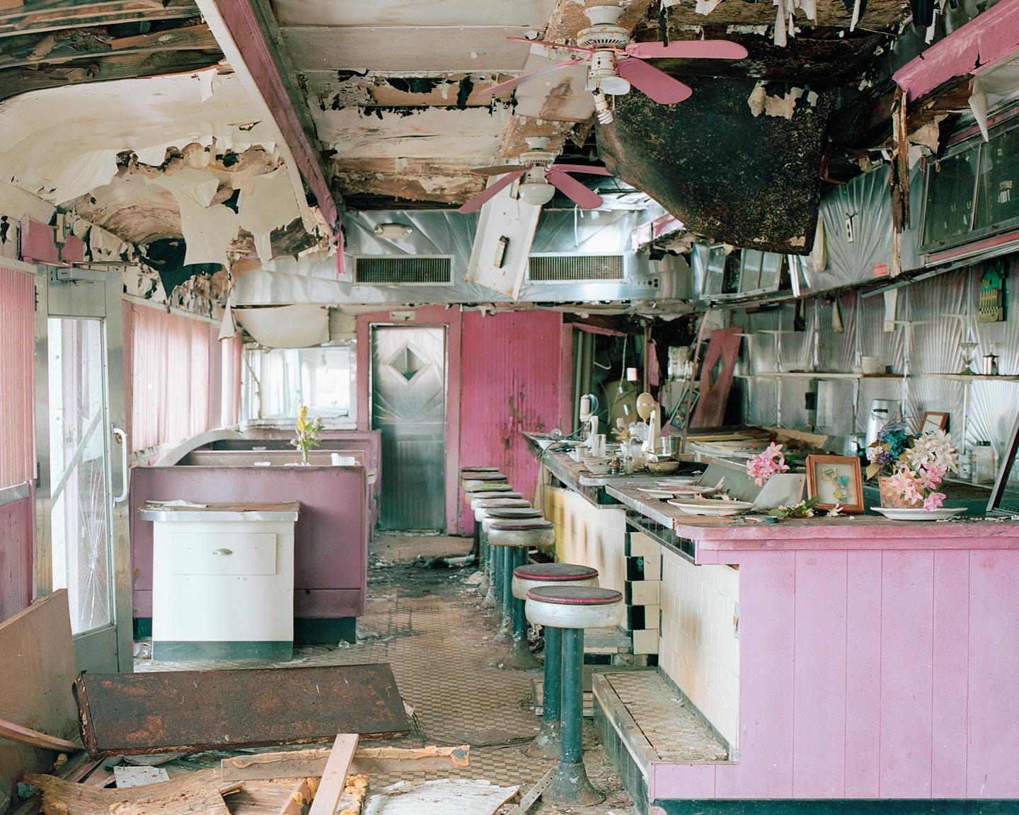 An abandoned diner with pink walls and booths has a damaged ceiling and debris on the floor.