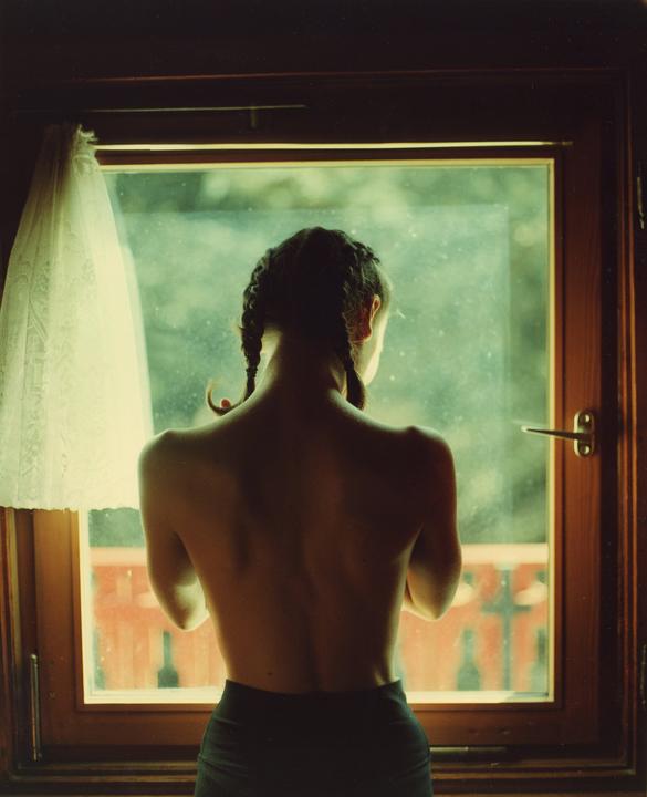 A woman with braided hair stands with her back to the viewer, looking out a window with a white curtain on the left.