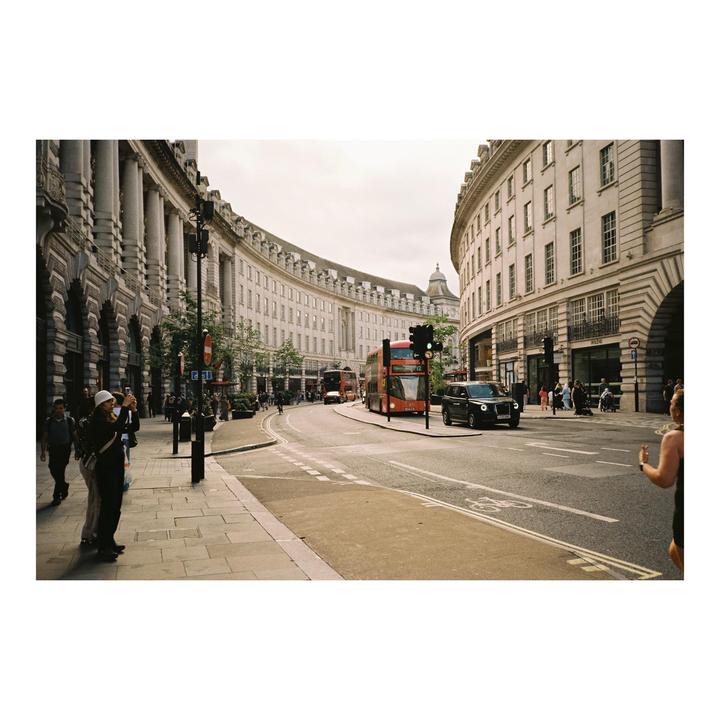 A street in London is filled with pedestrians, cars, and double-decker buses, flanked by large, curved buildings.