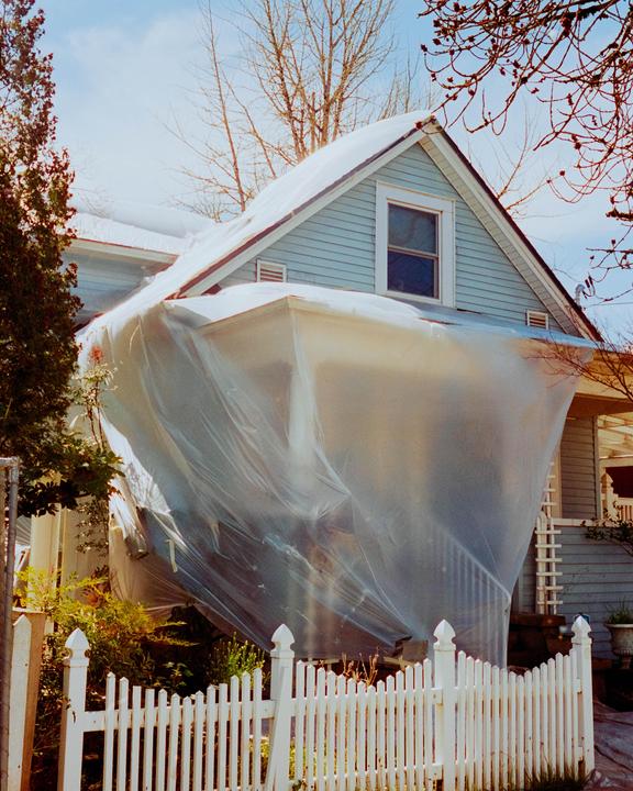 A light blue house is partially covered in clear plastic sheeting, with a white picket fence in the foreground.