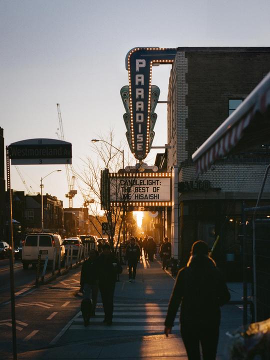 A street scene at sunset shows people walking on a sidewalk next to a street with cars and a building with a large sign that reads "Paradise" and a marquee that reads "Candlelight: The Best of Hisaishi."