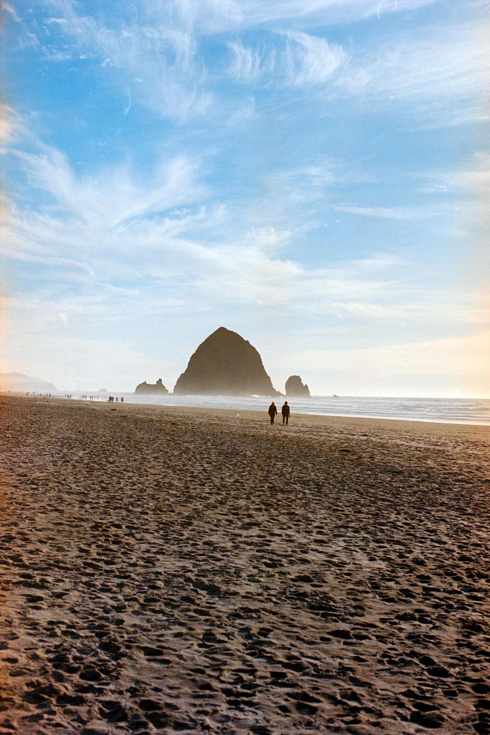 Two people walk on a sandy beach toward a large rock formation in the distance under a blue sky with wispy clouds.