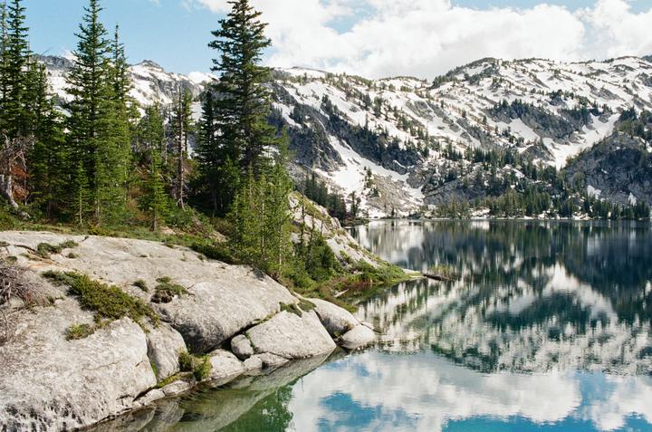 A calm lake reflects snow-capped mountains and a blue sky with clouds, surrounded by pine trees and rocks.