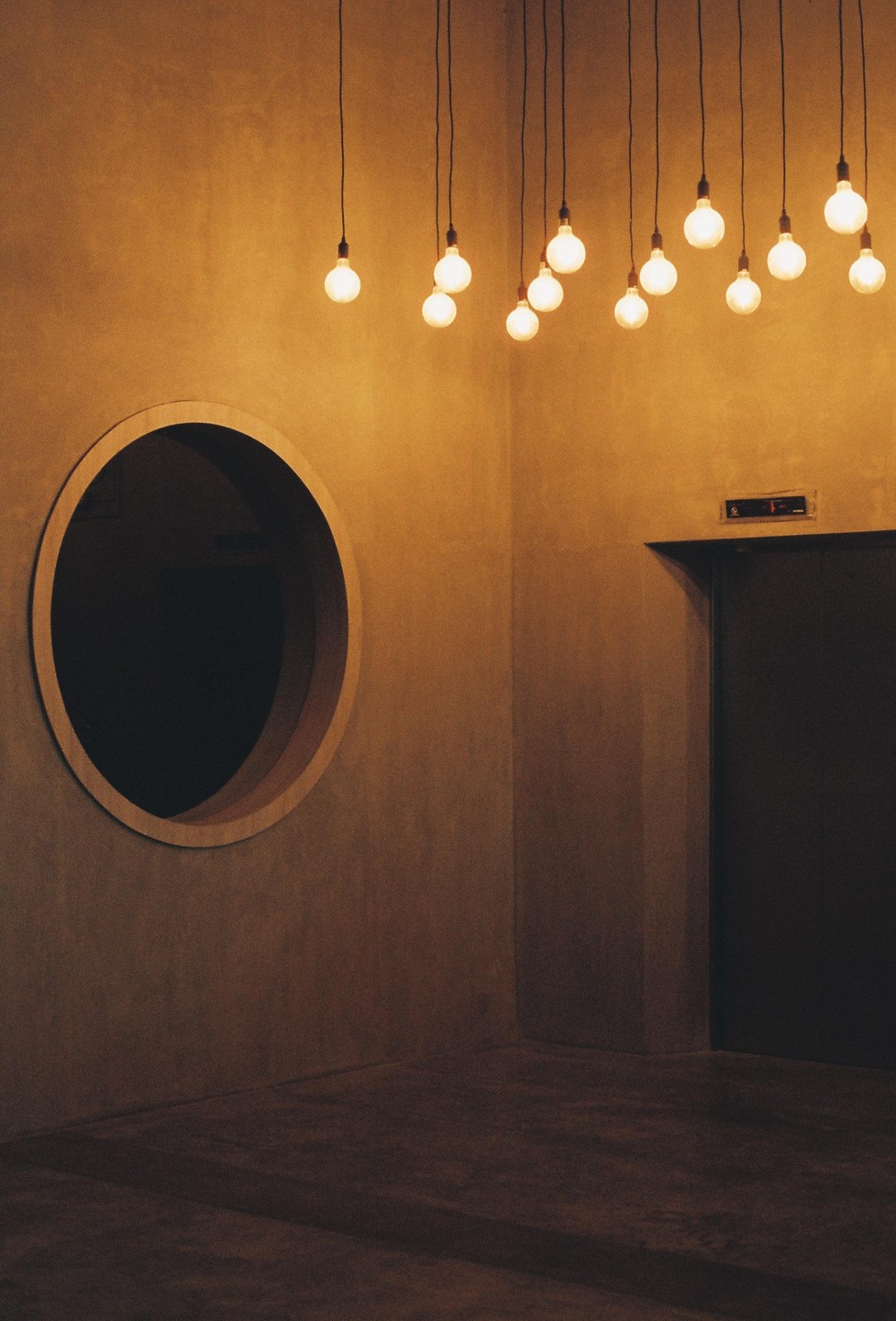 A corner of a room with a round window, an elevator door, and several hanging light bulbs.