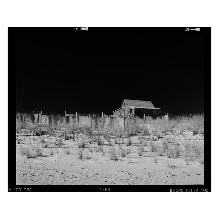 A small, weathered shack stands behind a wooden fence on a sandy beach with sparse vegetation under a dark sky.