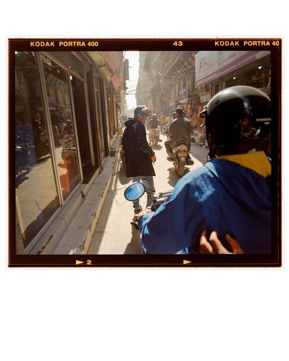 A man on a motorcycle looks back at the camera while another man walks past storefronts on a busy street.