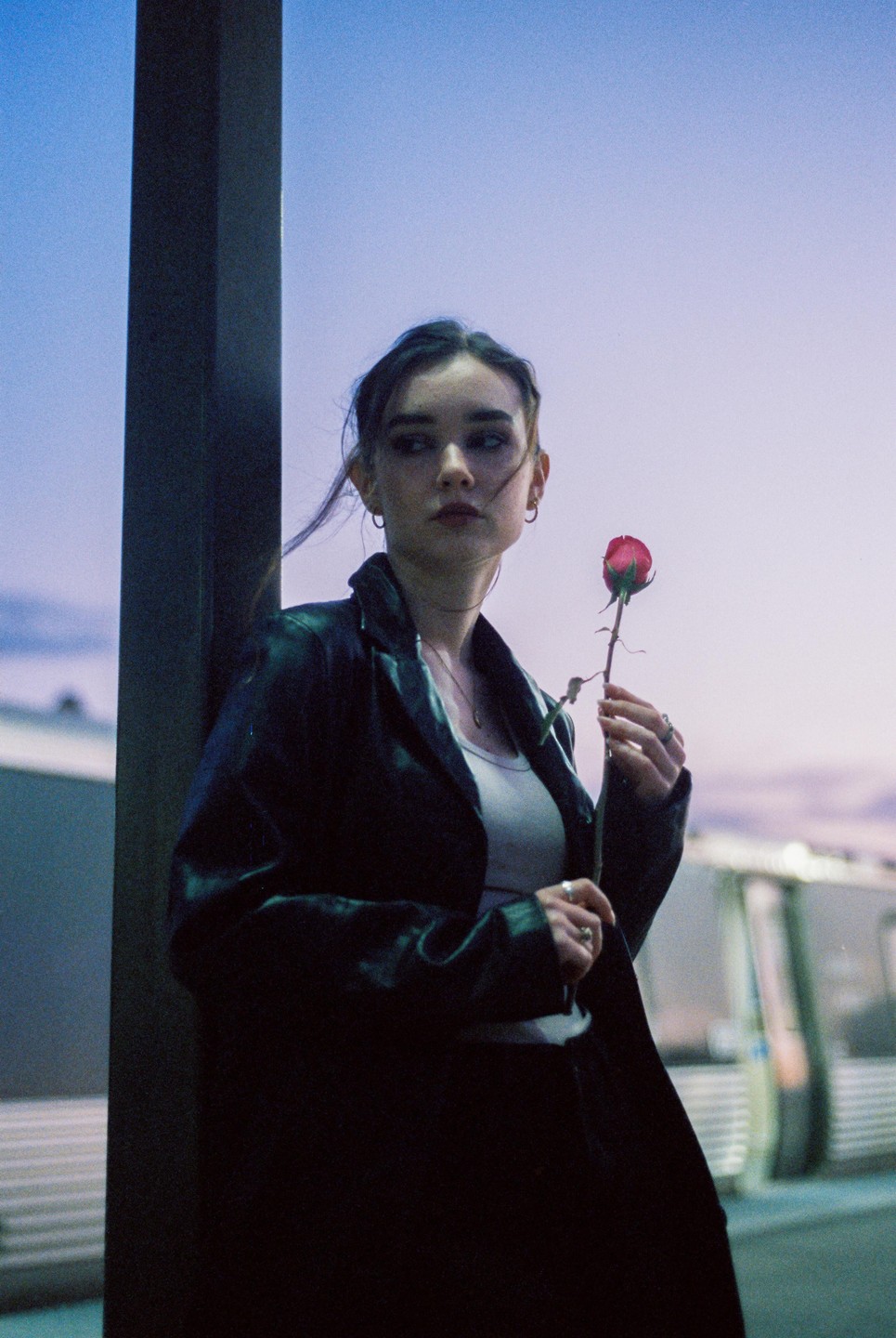 A woman in a black jacket holds a red rose at dusk.