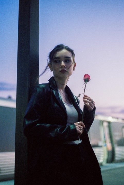 A woman in a black jacket holds a red rose at dusk.