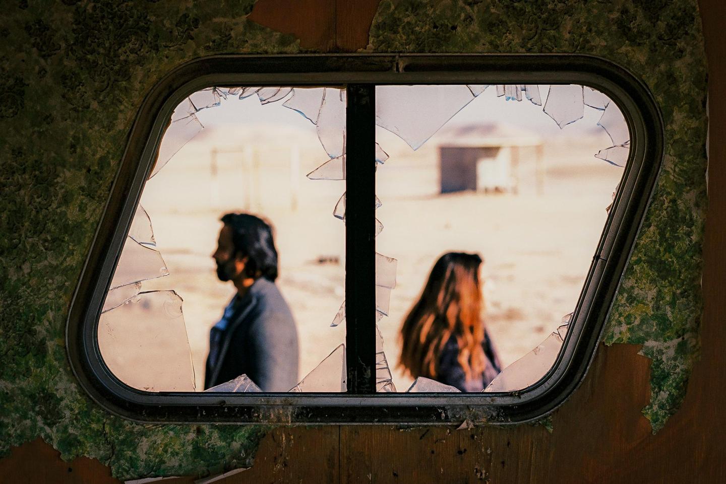A man and a woman are visible through a broken window with peeling wallpaper.