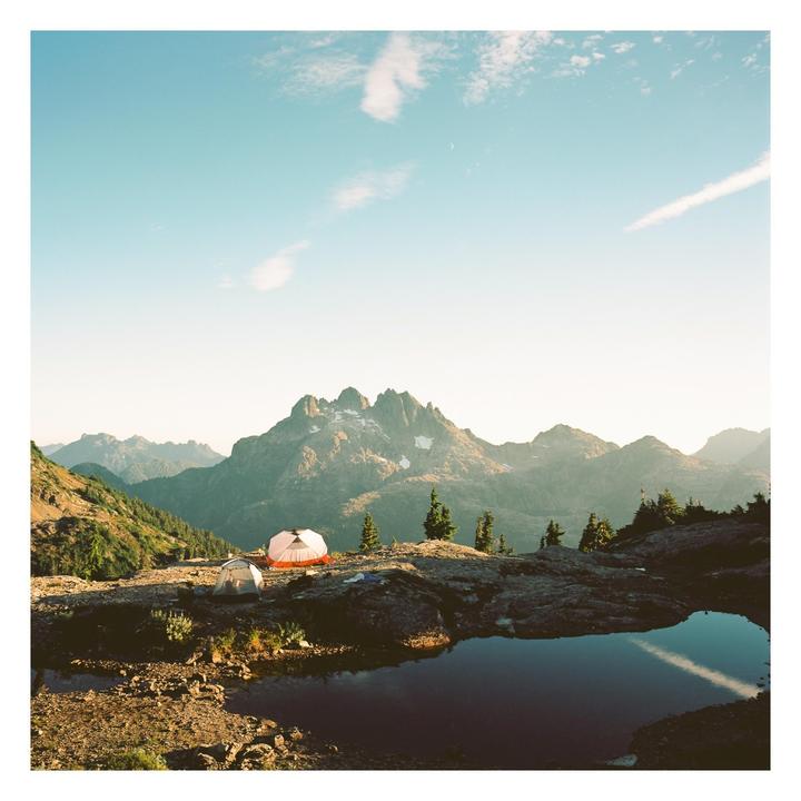 Two tents are pitched on a rocky mountain ledge overlooking a lake and distant peaks.
