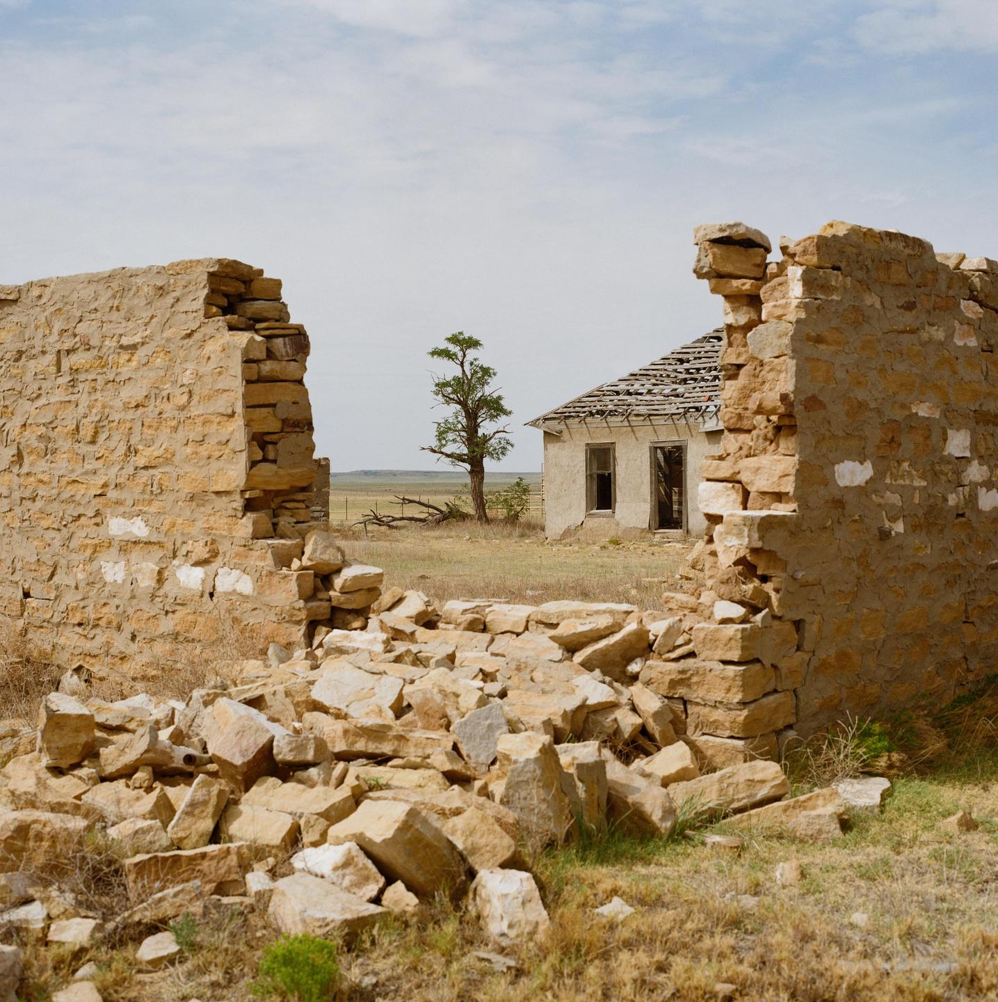 A dilapidated stone wall frames a view of a distant abandoned house and a lone tree in a dry, grassy field under a cloudy sky.