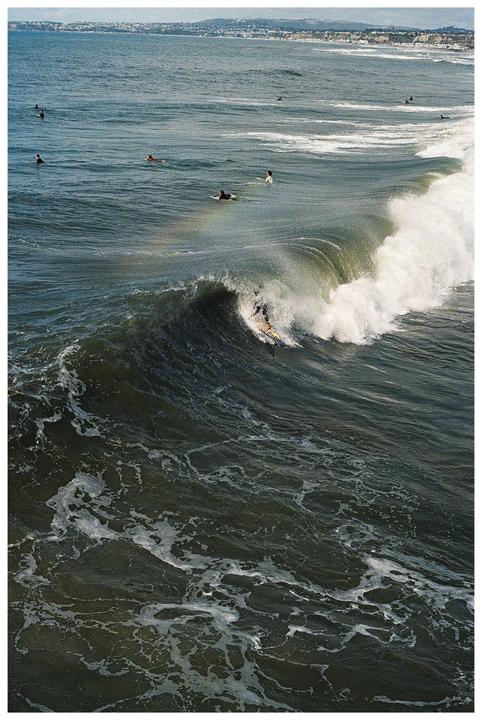 A surfer rides a large wave while other surfers wait in the water with a distant coastline visible.