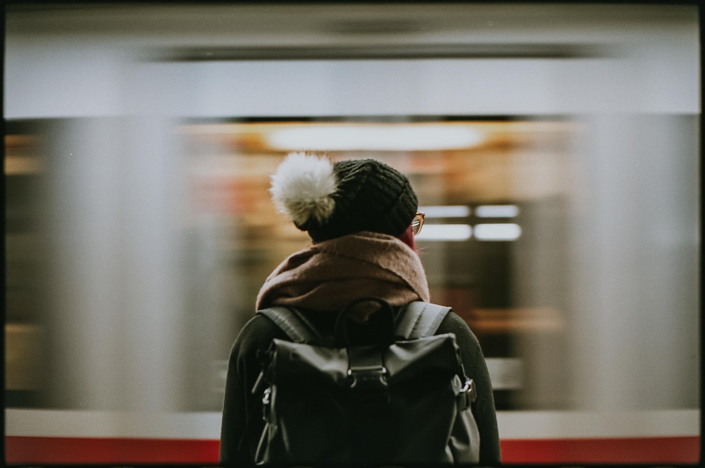 A person wearing a knit hat with a pom-pom and a backpack stands on a subway platform as a train passes by.