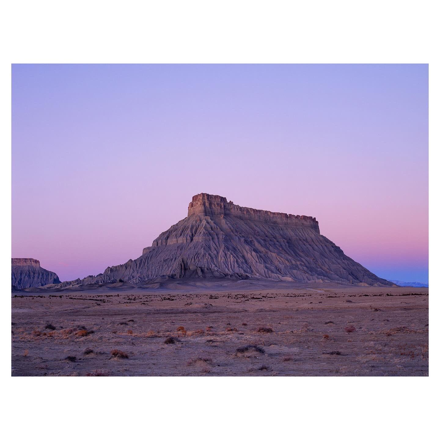 A large mesa rises from a flat, arid landscape under a purple and pink sky.