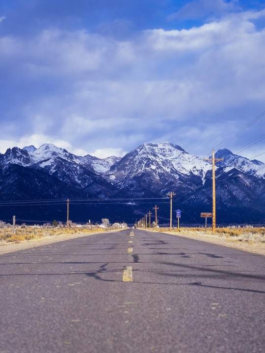 A long road leads towards snow-capped mountains under a cloudy sky.