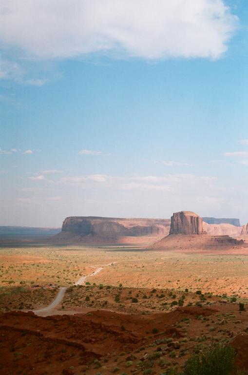 A dirt road leads through a vast desert landscape towards large rock formations under a blue sky with scattered clouds.