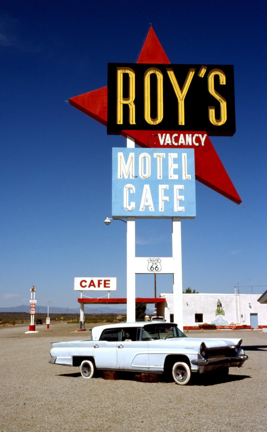 A light blue vintage car is parked in front of a motel and cafe sign with a red star and the words "Roy's Motel Cafe" and "Route 66" visible.