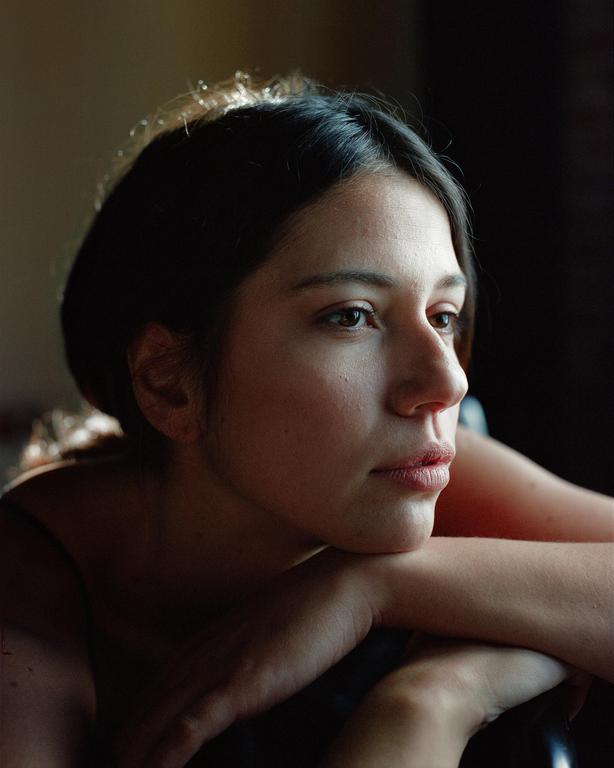 A woman with dark hair rests her chin on her crossed arms, looking to the right of the frame.