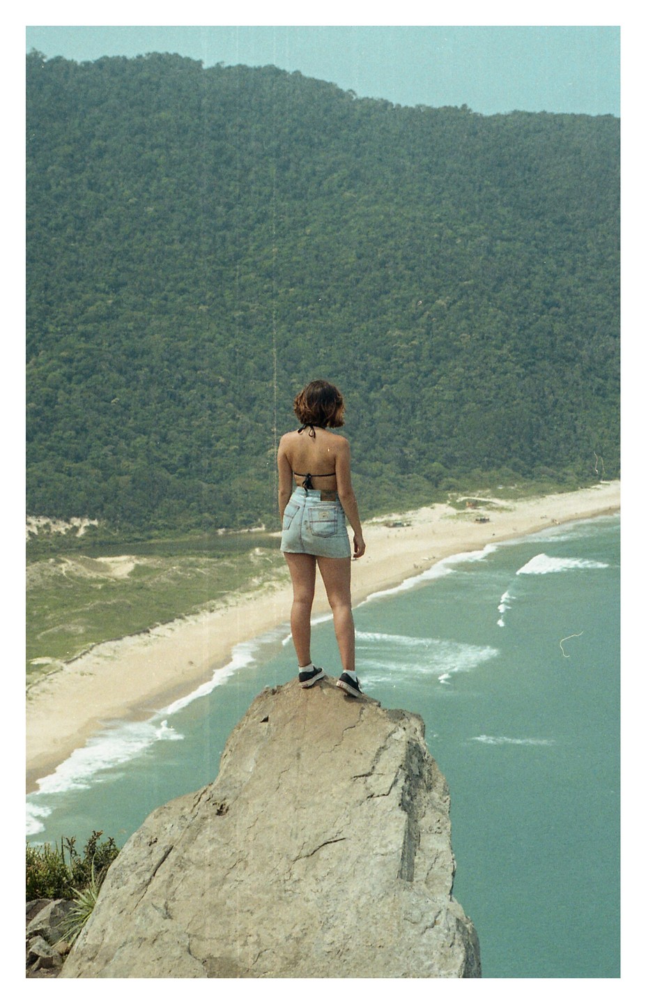 A woman stands on a rock overlooking a beach and ocean with a mountain in the background.