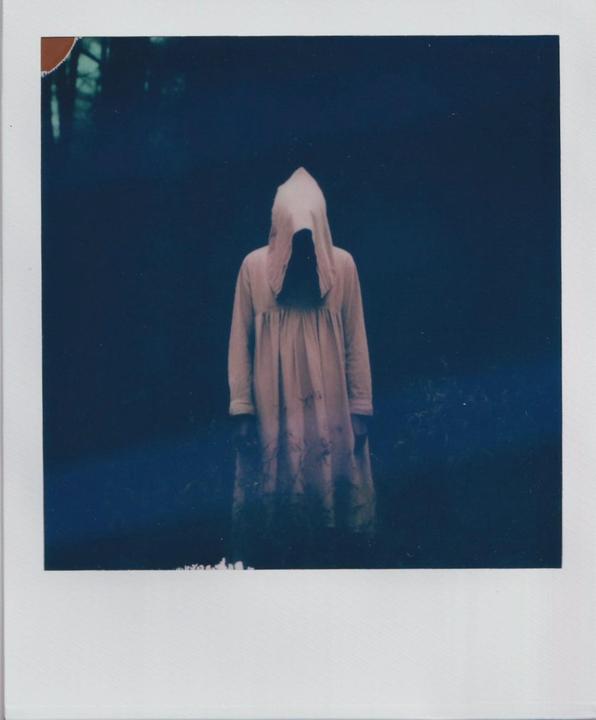 A figure in a light-colored hooded robe stands in a dark, shadowy environment.