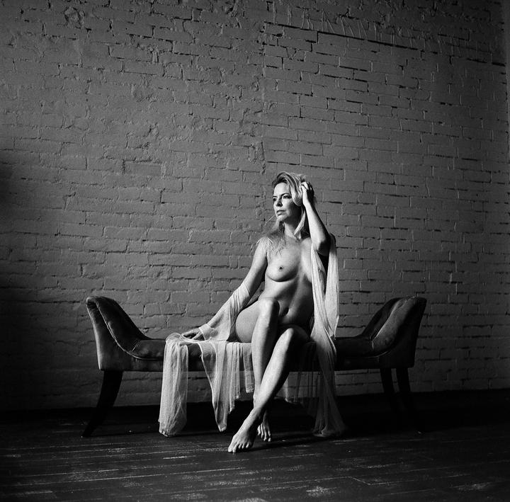 A woman with long hair sits on a bench in front of a brick wall, with a sheer fabric draped around her.